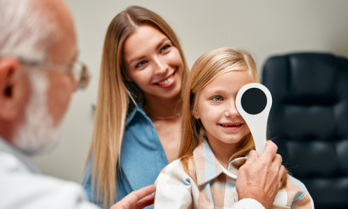 Cute little girl closing one eye during an ophthalmologist consultation while sitting on her mother's lap. A child points to an eye chart during an eye test.