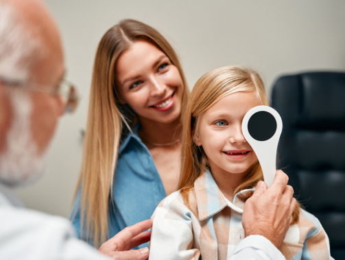Cute little girl closing one eye during an ophthalmologist consultation while sitting on her mother's lap. A child points to an eye chart during an eye test.